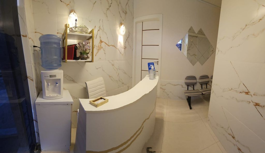 Área de recepção moderna da clínica odontológica Dra. Amanda Magre no Alto da Boa Vista, RJ, com balcão branco curvo, paredes de mármore, cantinho do café e ambiente iluminado.